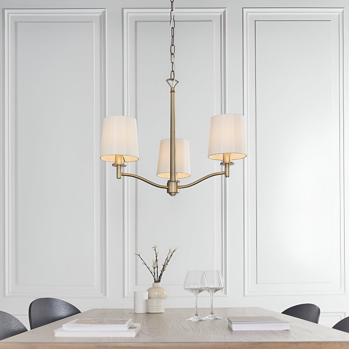 Endon Castwell 3 light pendant in antique brass with vintage white faux silk shades, hung above a dining table against panelled white walls.