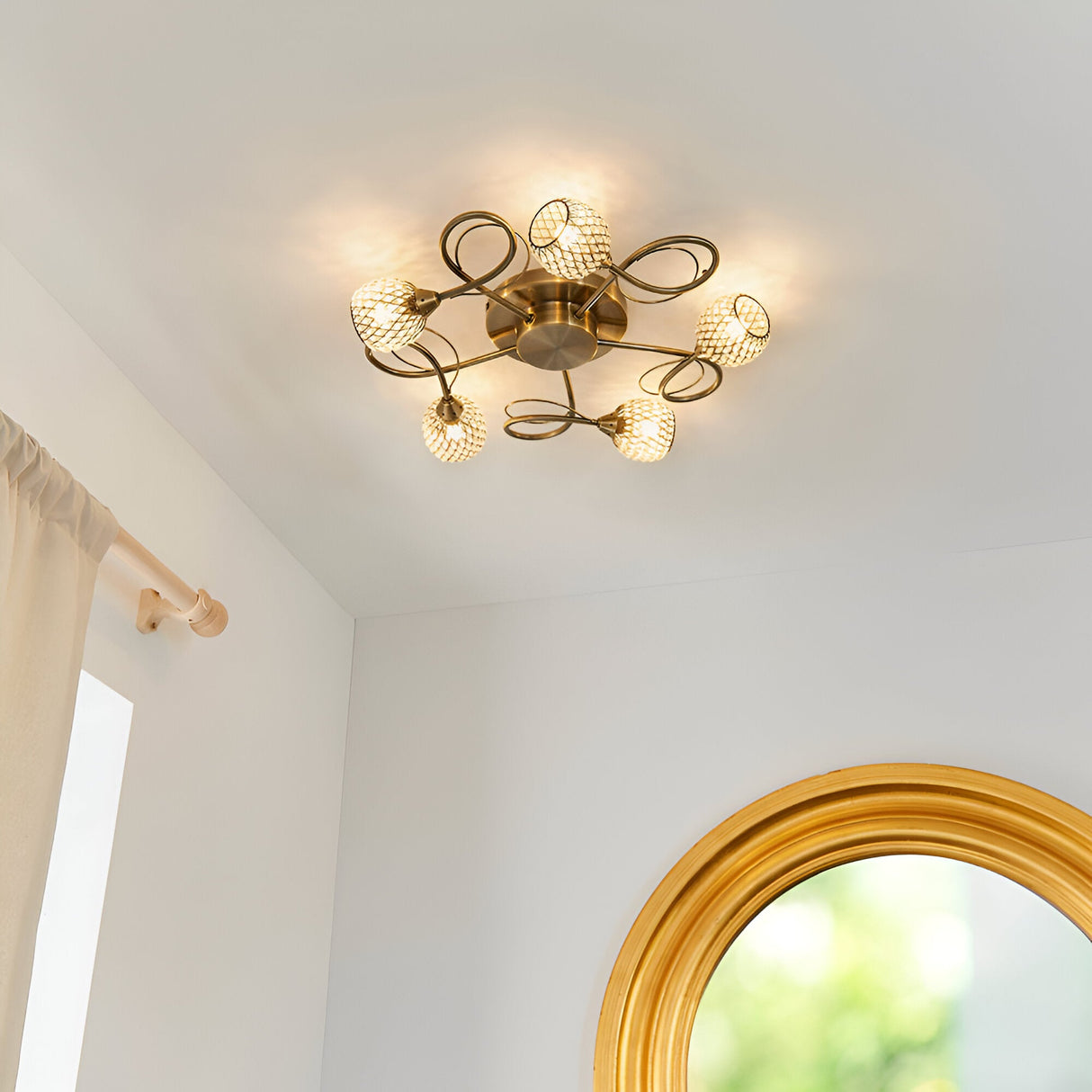 Endon Harcourt 5 light semi-flush ceiling light in antique brass with looped arms and textured glass globes, shown lit in a bright room near a gold mirror.