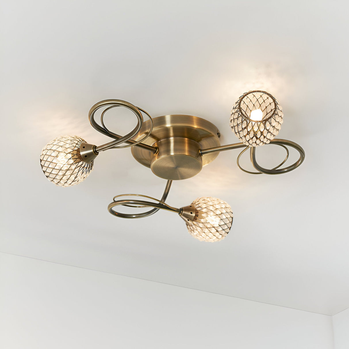 Endon Harcourt 3 light semi-flush ceiling light in antique brass with mesh-caged glass shades on curled arms, mounted to a white ceiling.
