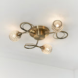 Endon Harcourt 3 light semi-flush ceiling light in antique brass with mesh-caged glass shades on curled arms, mounted to a white ceiling.