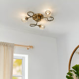 Endon Harcourt 3 light semi-flush ceiling light in antique brass with mesh glass shades on scrolled arms, installed on a white ceiling near a window.
