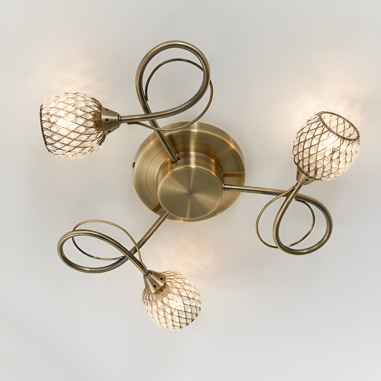 Endon Harcourt 3 light semi-flush ceiling light in antique brass, viewed from below showing curled arms and mesh-caged glass shades around a central disc.