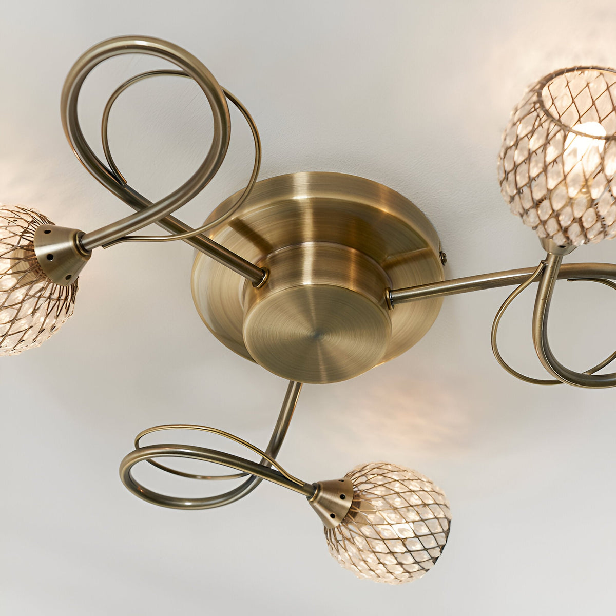Close-up of the Endon Harcourt 3 light semi-flush ceiling light in antique brass, showing the central fitting, looped arms, and mesh crystal shades.