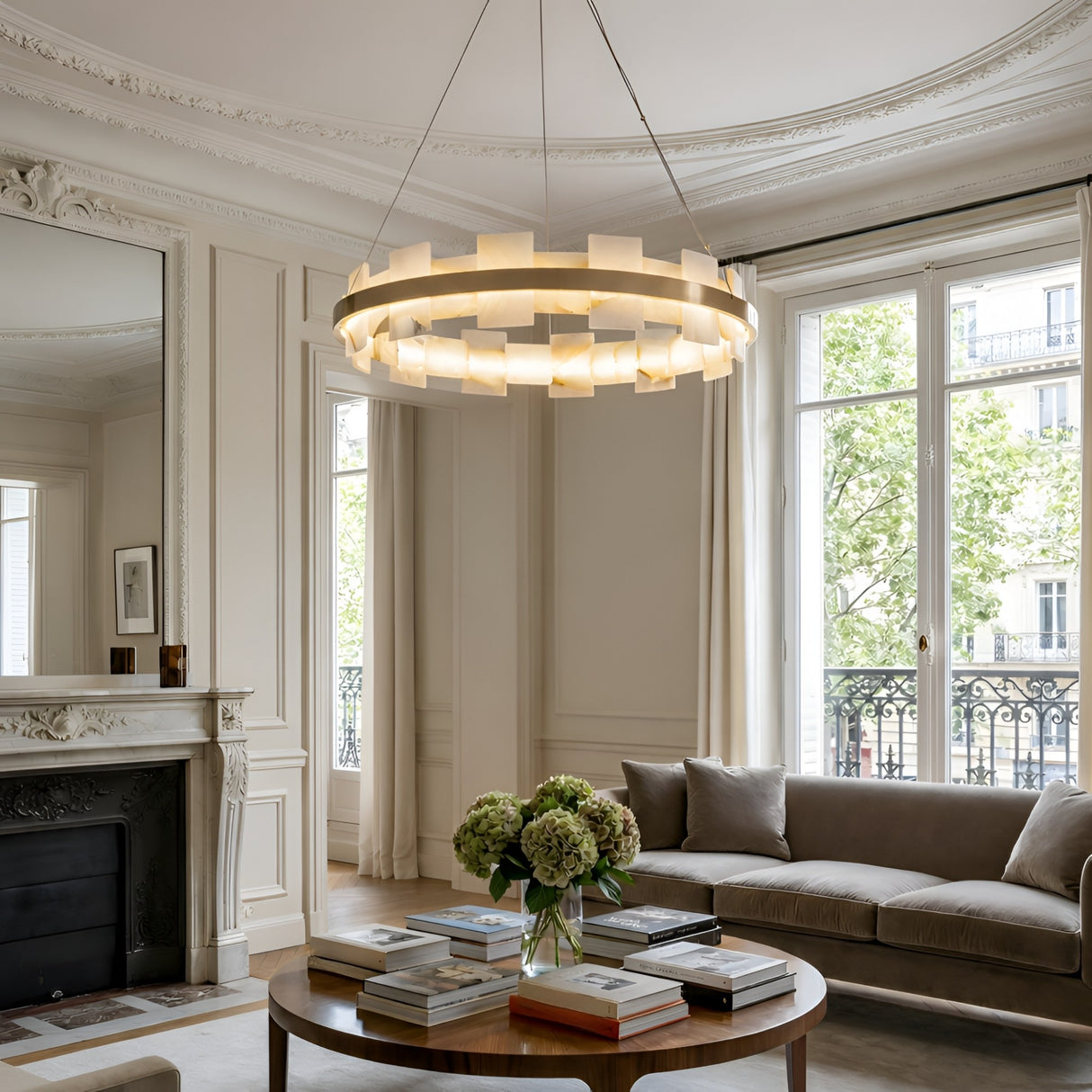 Elegant living room with ornate molding, a marble fireplace, large windows with sheer curtains, a Montyra LED Small Panelled Alabaster Chandelier in Soft White & Brass, a round coffee table with books and flowers, and a beige sofa.