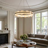 Elegant living room with ornate molding, a marble fireplace, large windows with sheer curtains, a Montyra LED Small Panelled Alabaster Chandelier in Soft White & Brass, a round coffee table with books and flowers, and a beige sofa.