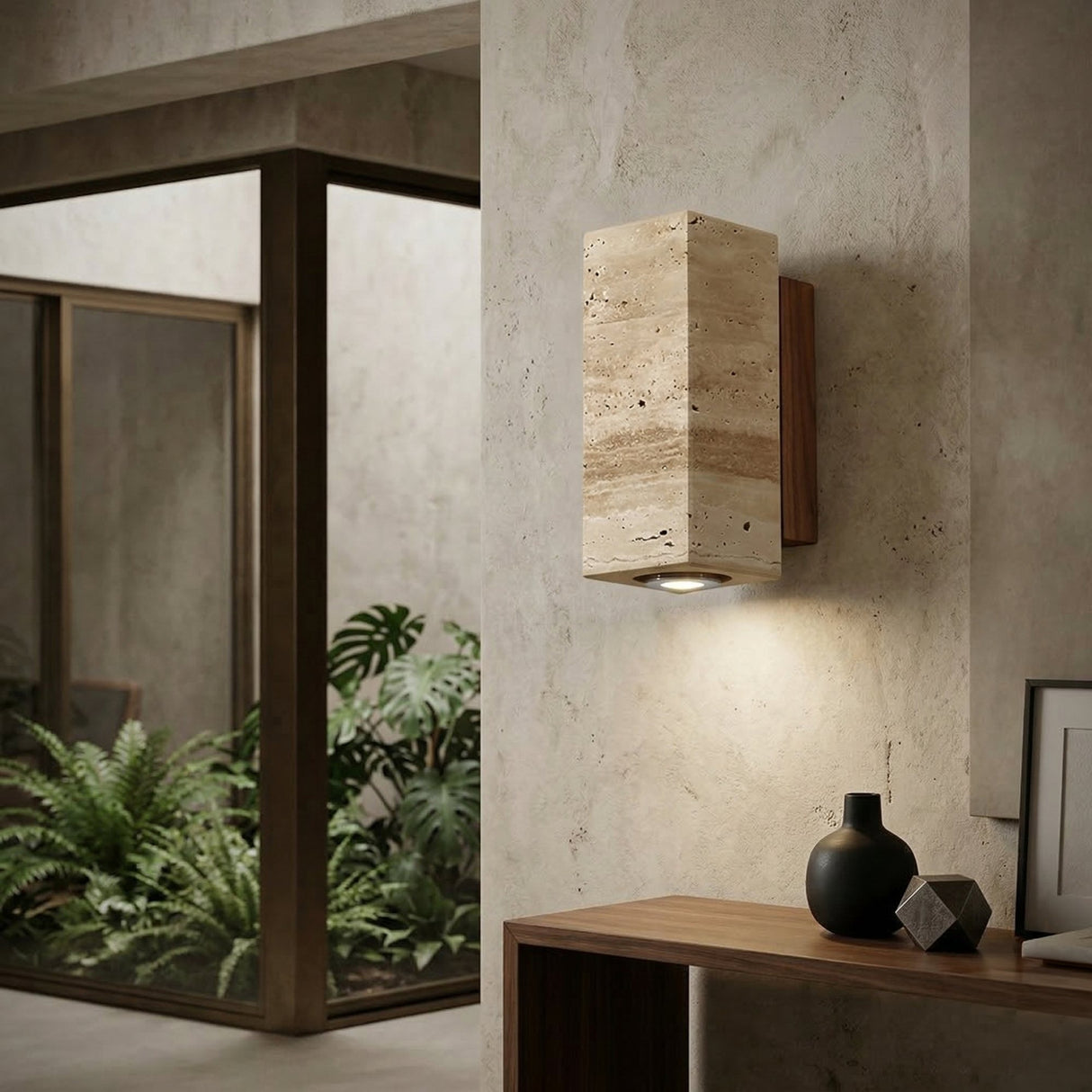 The Bravora 1 Light Small Rectangle Travertine Wall Light in Walnut Brown & Light Beige is mounted on a textured beige wall near a wooden console with minimalist decor, as large windows look out onto lush green plants.