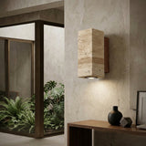The Bravora 1 Light Small Rectangle Travertine Wall Light in Walnut Brown & Light Beige is mounted on a textured beige wall near a wooden console with minimalist decor, as large windows look out onto lush green plants.