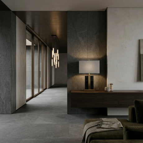 A modern living space with dark stone floors and walls, a floating console topped with the Elvaris 1 Light Framed Marble Table Lamp in brass & black, pendant lights in the hallway, glass doors, and a partially visible green armchair.