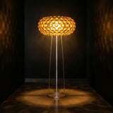 The Briller LED Floor Light - Gold features a circular honeycomb-patterned shade that emits warm, ambient light and casts intricate shadows across dark walls and floors.
