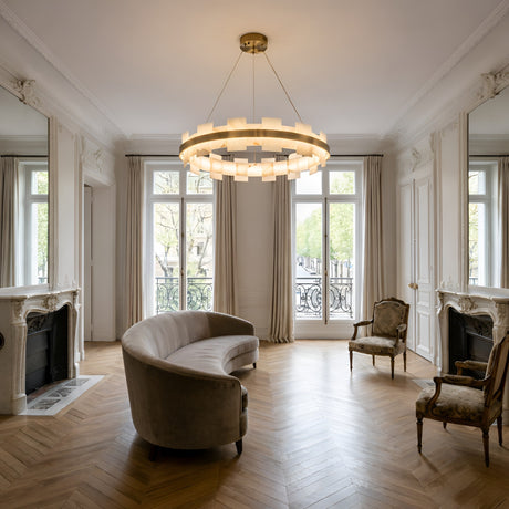 Elegant living room with herringbone wood floors, large windows, a Montyra LED Large Panelled Alabaster Chandelier in soft white & brass, a curved sofa, two antique armchairs, white walls, ornate fireplaces, and classic molding.