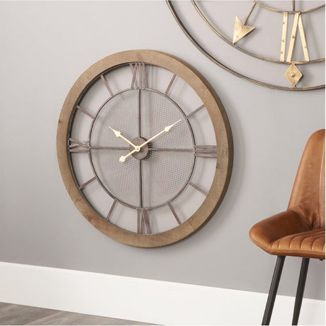 Round 80cm wall clock with wood frame, black Roman numerals, iron mesh centre, and gold hands, shown above a chair beside a partial gold wall art piece.