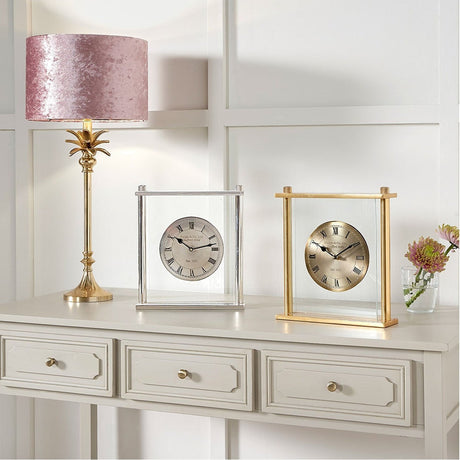 Gold brass and glass mantel clock with floating clock face, 28cm, shown beside a matching silver-framed clock on a console table with a pink lamp.