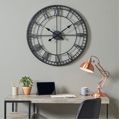 Black Iron Round Large Wall Clock, 80cm, with skeleton dial and Roman numerals, hung above a home office desk with a copper lamp and tablet.