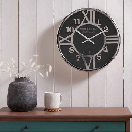 Silver Iron & Black Face Round Wall Clock, 40cm, with Roman and Arabic numerals, silver hands, shown mounted on a white panelled wall above a wooden sideboard.