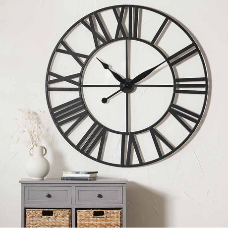 Black Iron Large Round Wall Clock, 120cm, with skeleton Roman numerals and black hands, shown above a grey drawer unit with wicker baskets and a white vase.