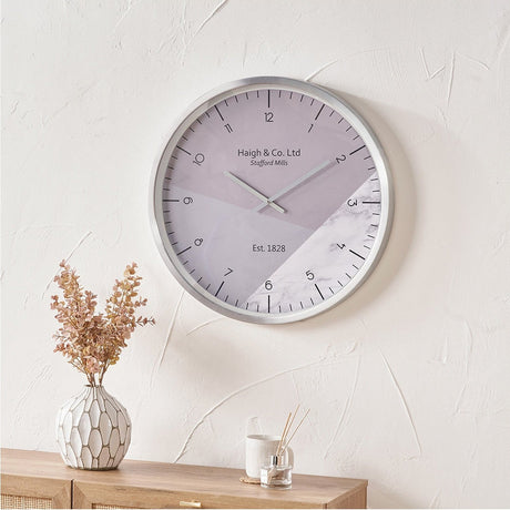 Silver aluminium 60cm wall clock with grey and marble-effect printed face, displayed on a white wall above a wooden sideboard.