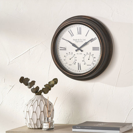 Antique bronze effect indoor outdoor wall clock, 42cm, with white Roman numeral dial, humidity and temperature subdials, mounted on a light wall above a console table.