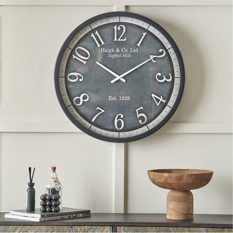 Round wall clock with black frame and dark grey face, oversized white numerals, "Haigh & Co. Ltd" text, shown mounted above a sideboard with decor accessories.