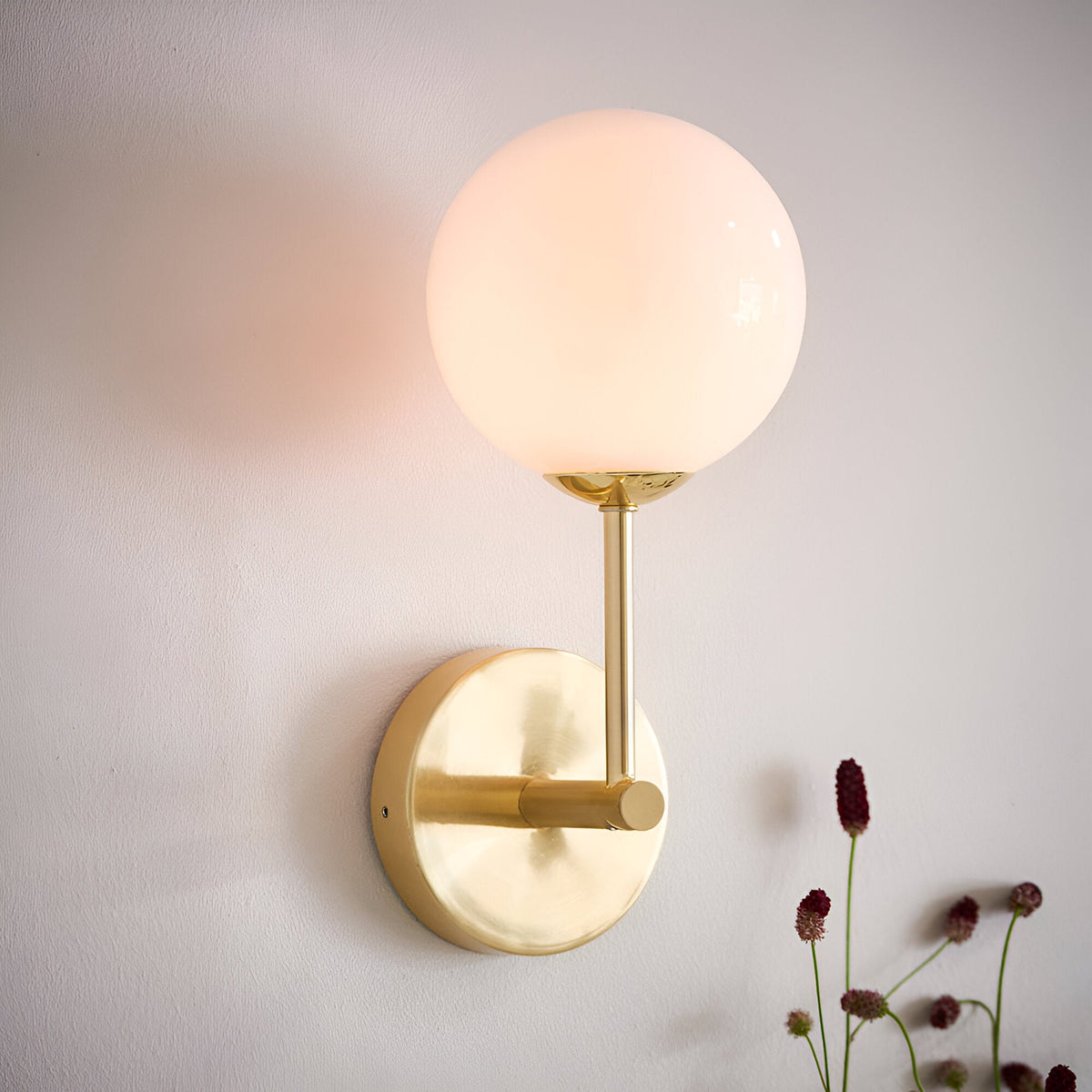 Endon Sinclair wall light with a gloss opal glass globe shade on a brushed brass stem and round backplate, mounted on a pale wall beside dried flowers.