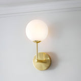 Endon Sinclair wall light with a single opal glass globe on a slim brushed brass stem and round backplate, mounted on a white wall below cornice moulding.