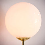 Endon Sinclair wall light illuminated, showing its round gloss opal glass globe shade seated on a brushed brass fitting against a pale wall.
