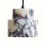 A close-up of the Valmere 1 Light Veined Marble Single Pendant Light in white and black, featuring a cylindrical, tiered white veined marble shade suspended by a black cord against a plain background.
