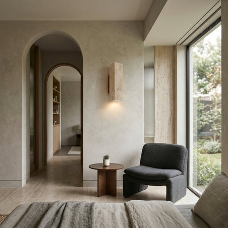 A modern, minimalist living space with a dark gray armchair, walnut brown round side table with a potted plant, large garden-view window, light-toned walls, and a Bravora 1 Light Large Rectangle Travertine Wall Light in walnut brown & light beige by an arched doorway.