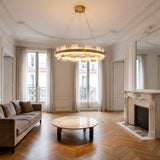 A spacious, elegant living room with a Montyra LED Small Panelled Alabaster Chandelier in Soft White & Brass, a brown sofa, round marble coffee table, ornate white fireplace, herringbone wood floor, tall windows, light curtains, and ceiling moldings.
