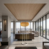 A modern, spacious living room features floor-to-ceiling windows with city views, neutral-tone furniture, a sculptural light fixture, the Briller LED Floor Light in gold, and a wood-paneled ceiling for a luxurious atmosphere and warm ambient lighting.