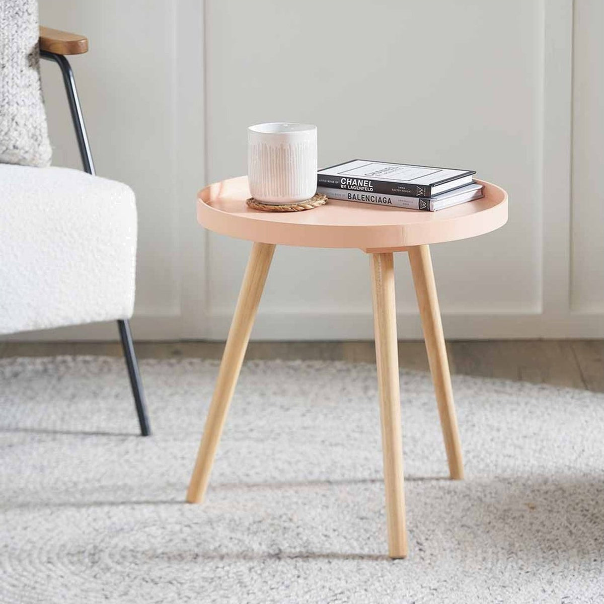 Halston round side table with apricot wood veneer top and pine tripod legs, styled with a mug and books beside an armchair in a living room.