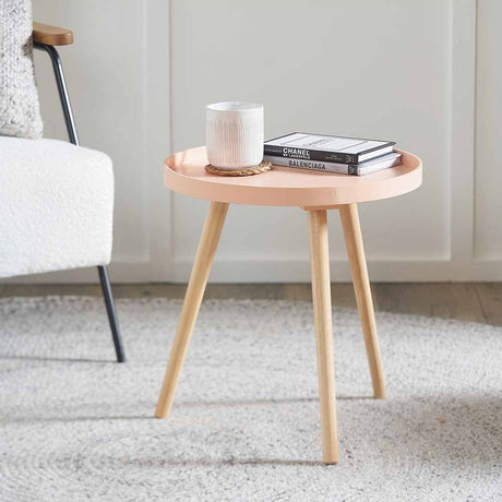 Halston round side table with apricot wood veneer top and pine tripod legs, styled with a mug and books beside an armchair in a living room.