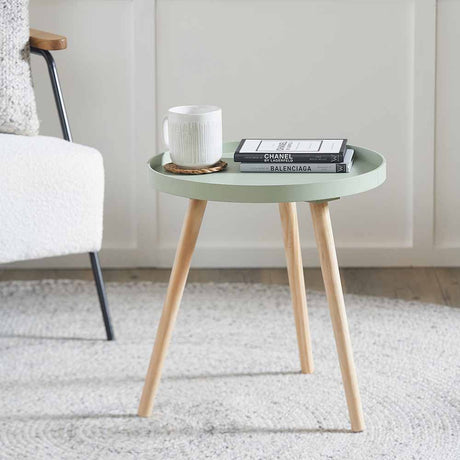 Halston Sage Green Wood Veneer & Pine Round Side Table, 41cm, with tapered wooden legs, styled with a mug and books beside an armchair on a living room rug.