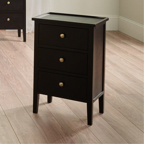 Chelmsford satin black pine 3 drawer unit with a raised-edge top and round brass knobs, shown in a bedroom setting on a wooden floor.