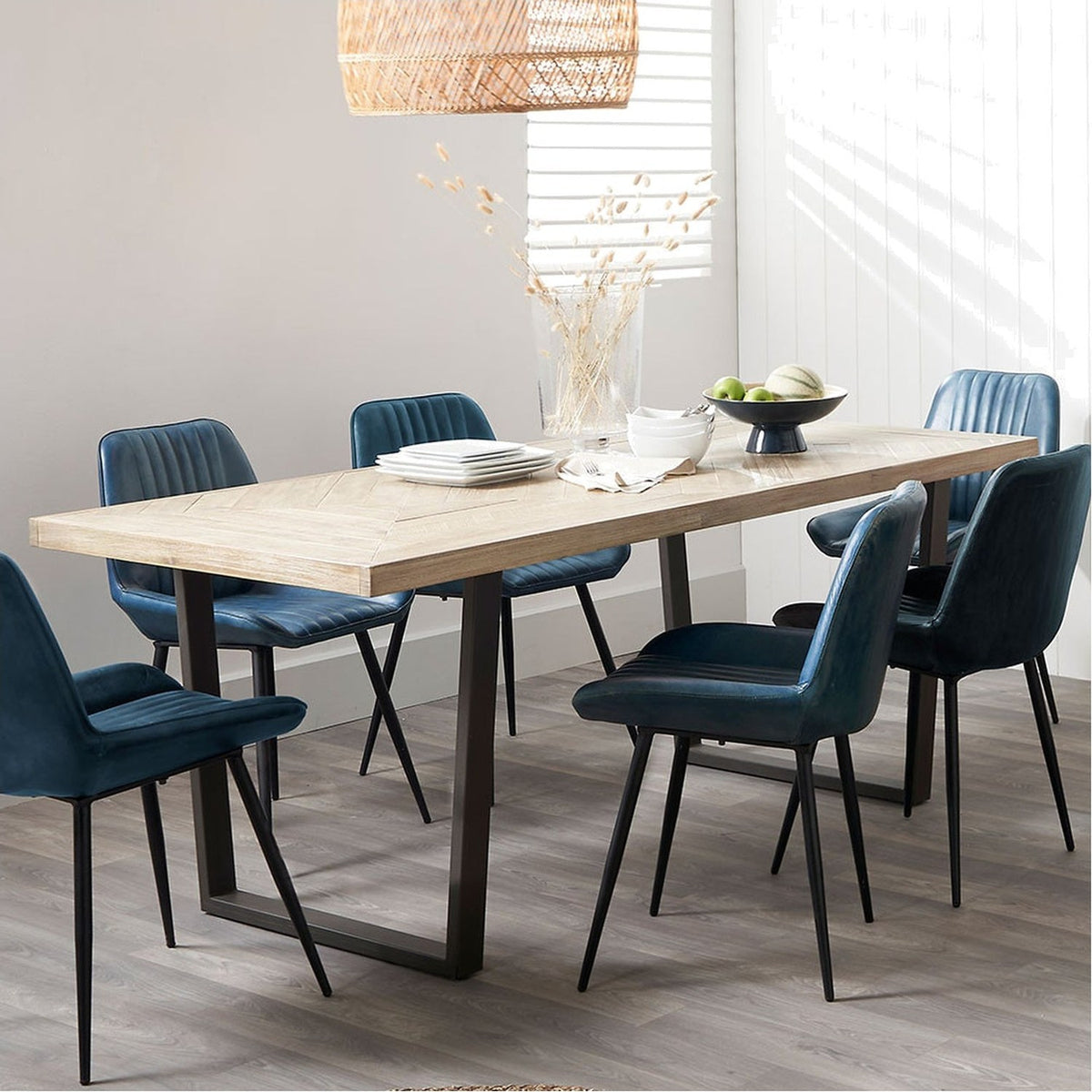 Marca dining table with a sand wash acacia wood herringbone-effect top and black iron U-shaped legs, styled with blue upholstered chairs in a dining room setting.