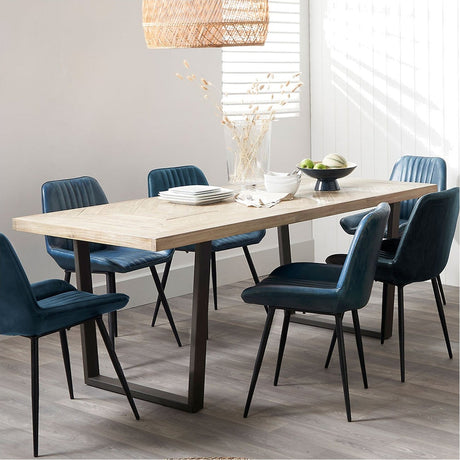 Marca dining table with a sand wash acacia wood herringbone-effect top and black iron U-shaped legs, styled with blue upholstered chairs in a dining room setting.