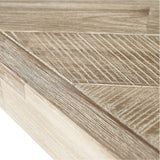 Close-up of the Marca dining table's corner, showing sand wash acacia wood with a chevron parquet pattern and visible grain texture in a light beige finish.