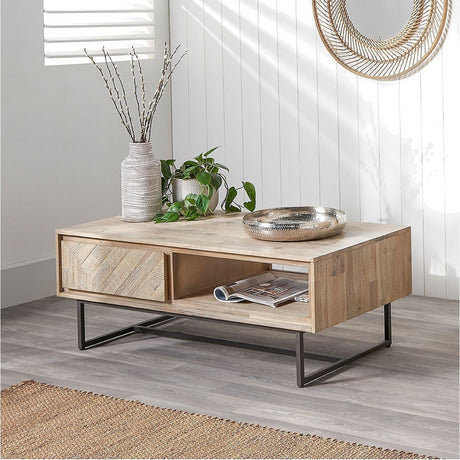 Marca sand wash acacia coffee table with two sliding herringbone-panel drawers, open shelf, and black iron frame, styled in a living room with decor accessories.