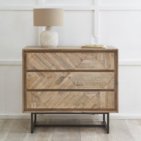 Marca 3 drawer chest in sand wash acacia wood with chevron parquet fronts and black iron base, styled with a lamp and books in a bedroom setting.