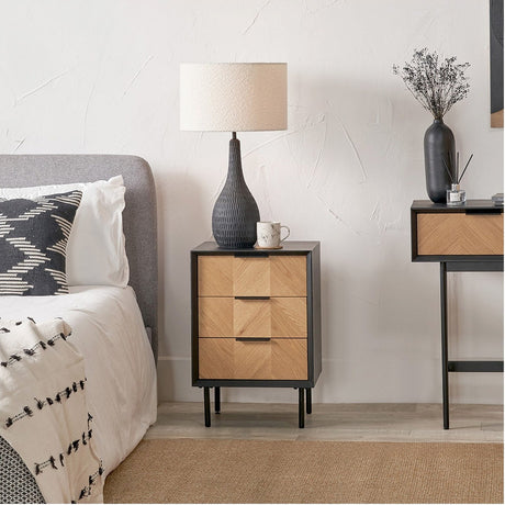 Hailee Black & Chevron Oak & Pine 3 Drawer Unit as a bedside table, black frame with chevron oak drawer fronts, black handles and tapered legs, shown beside a bed in a bedroom setting.