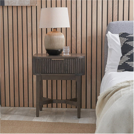 Raie Dark Brown Ribbed Oak & Pine 1 Drawer Unit used as a bedside table, with rounded top, ribbed front, brass handle, and splayed legs, styled beside a bed with a table lamp on top.