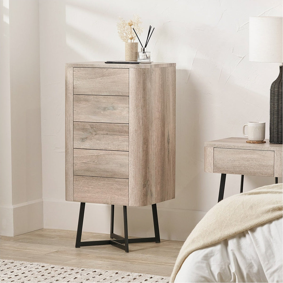 Ukiah tall chest of drawers in grey oak veneer with five drawers and angled black metal legs, shown in a bedroom beside a matching nightstand.