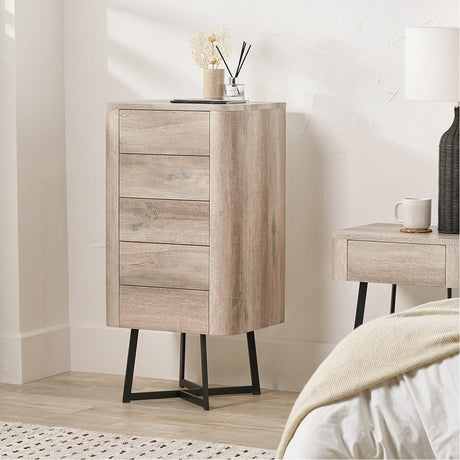 Ukiah tall chest of drawers in grey oak veneer with five drawers and angled black metal legs, shown in a bedroom beside a matching nightstand.