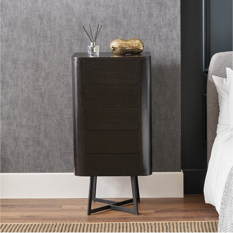 Ukiah black ash veneer 5 drawer tall chest with black metal cross-frame legs, shown in a bedroom beside a bed against a grey wall.