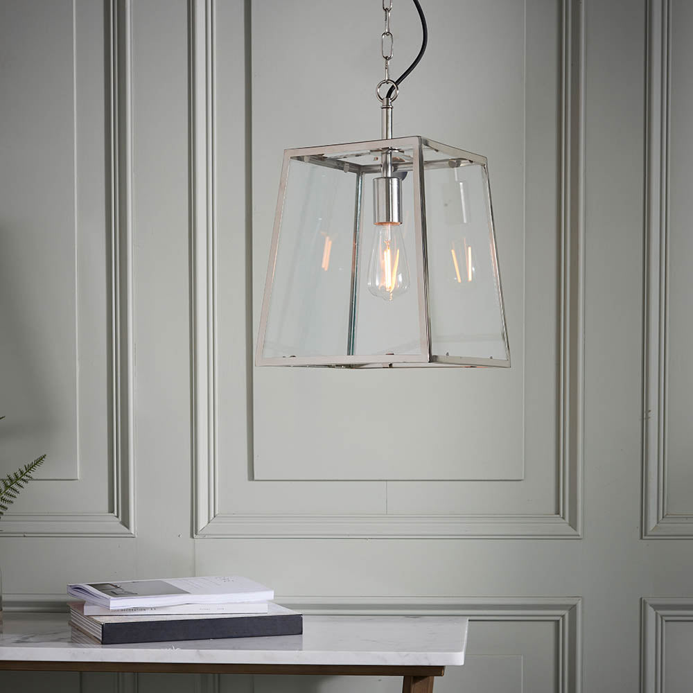 Brenton Lantern Pendant in nickel with clear glass panels and exposed filament bulb, hanging above a marble console table in a panelled room.