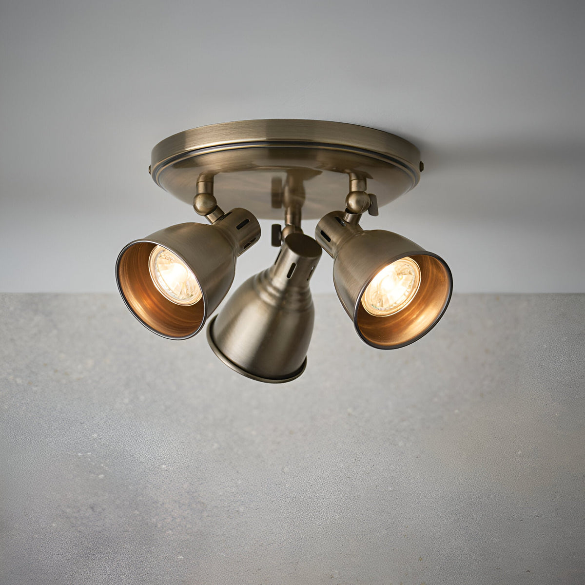 Endon Daneford 3 light round ceiling spotlight in antique brass with three independently adjustable steel heads, shown lit against a plain grey background.