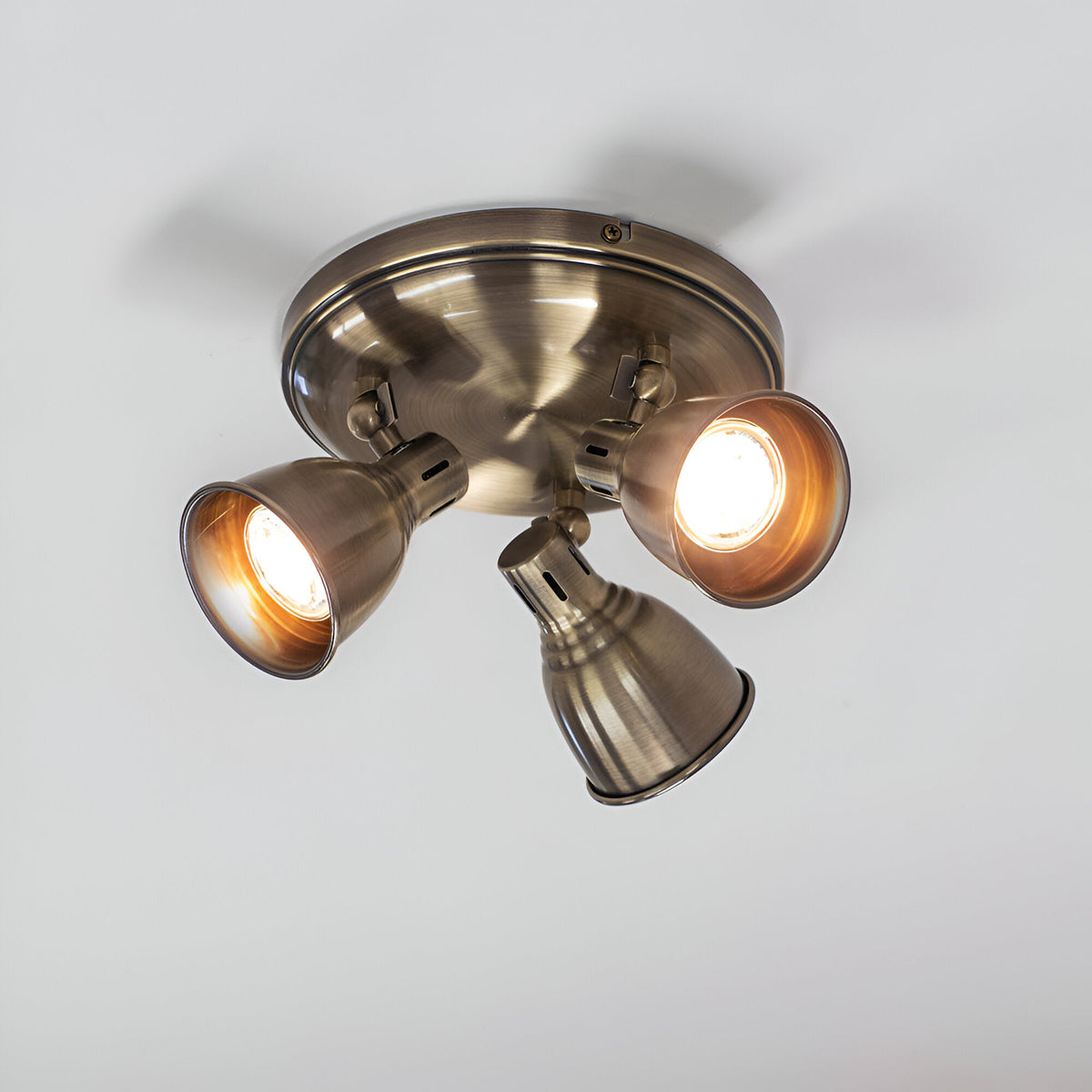 Endon Daneford 3 light round spotlight in antique brass, three adjustable heads on a circular ceiling plate, mounted on a white ceiling.