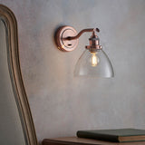 Endon Wentworth copper wall light with adjustable knuckle, clear glass shade and lit E14 filament bulb, mounted on a grey wall beside an upholstered chair.