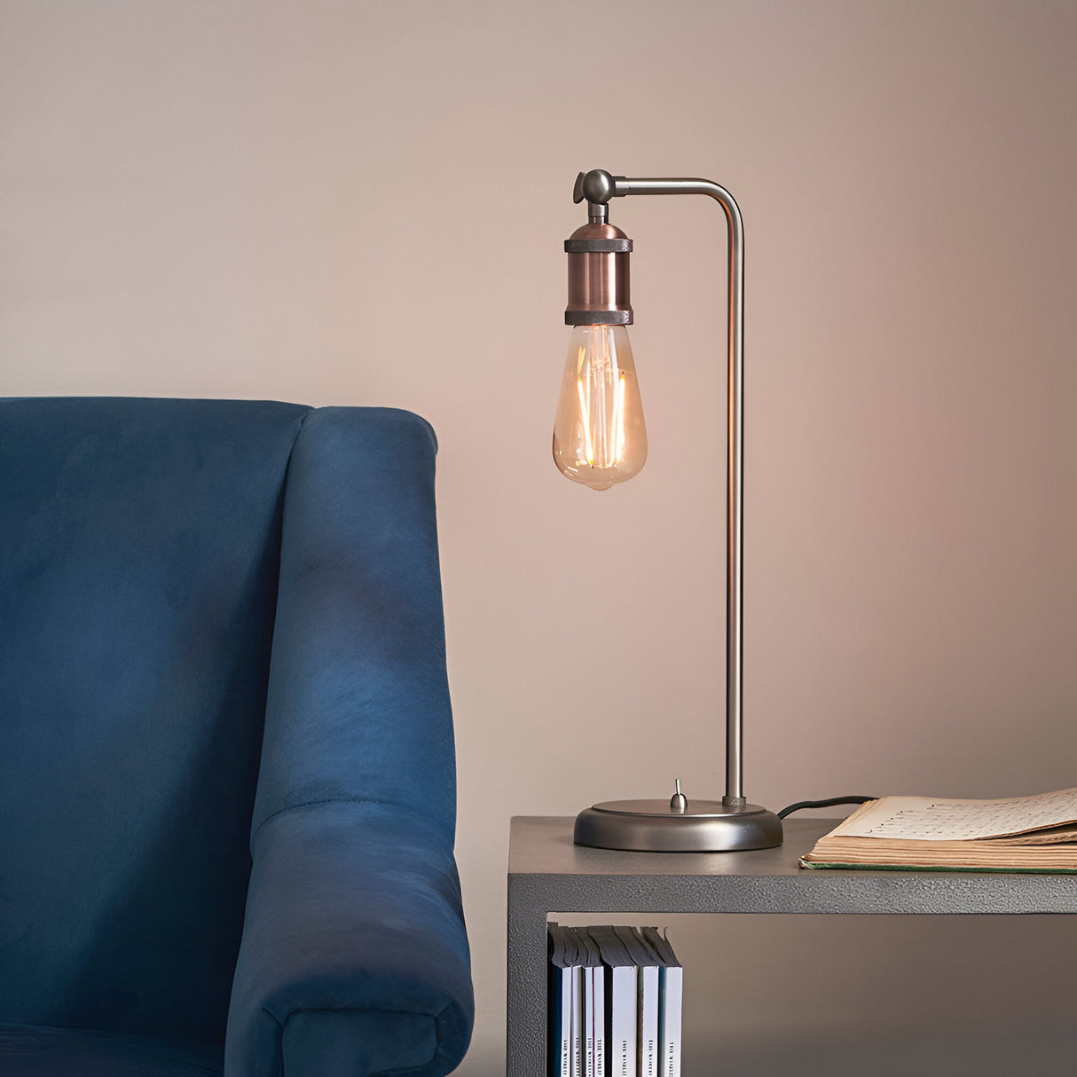 Brackley Table Lamp in copper and silver metal with an exposed E27 bulb, angled arm, and base switch, shown on a side table beside a blue armchair.