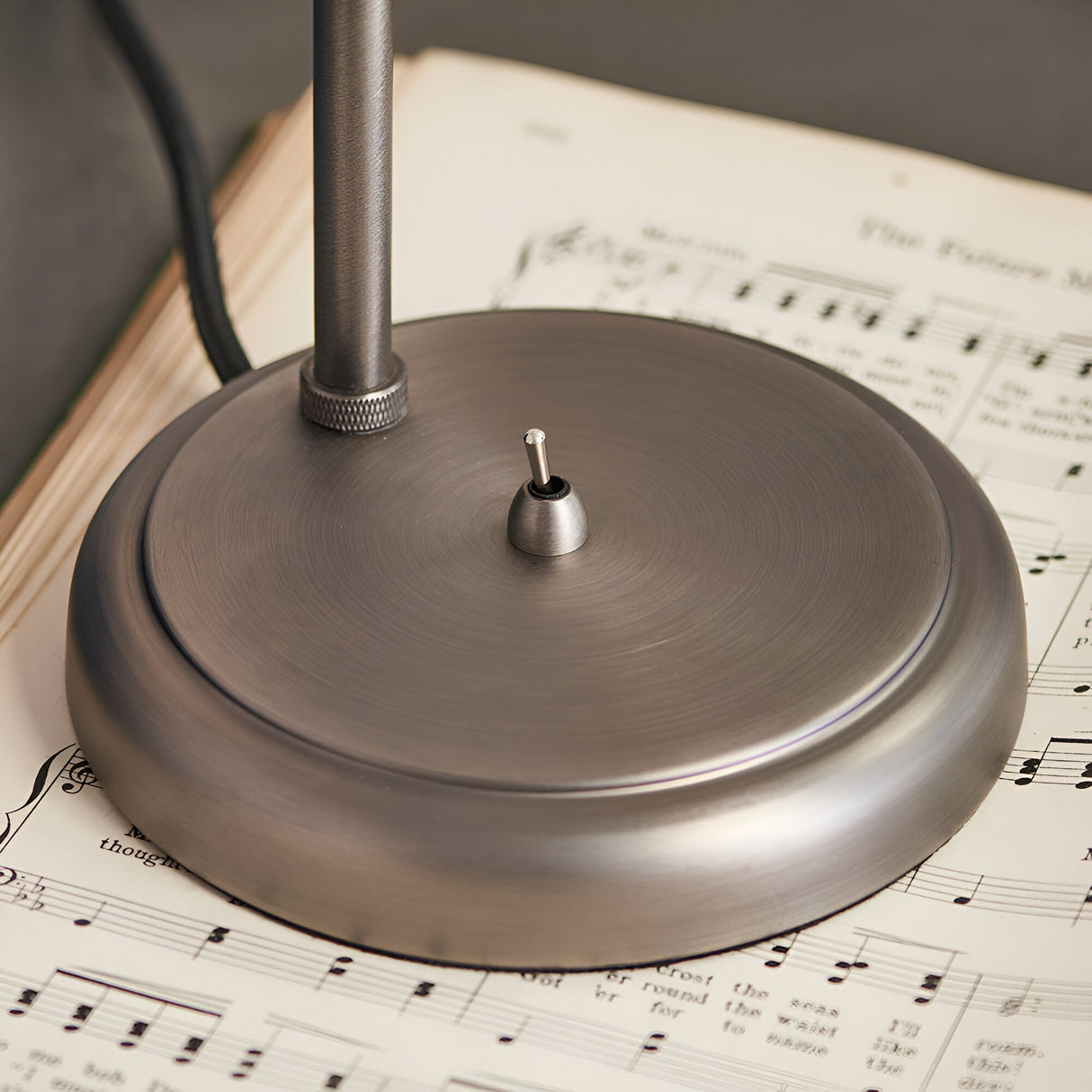 Close-up of the Brackley Table Lamp's round metal base in silver, showing the knurled adjustable joint and on/off toggle switch, set on sheet music.