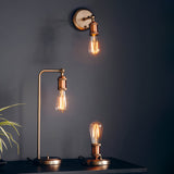 Brackley Table Lamp in copper with silver base, adjustable industrial-style arm and exposed E27 bulb, shown lit on a dark shelf beside a matching wall light.